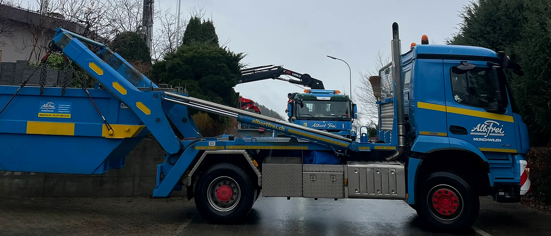Muldenlastwagen von Alb. Frei stellt eine Welaki-Mulde ab, dahinter ein Kranlastwagen